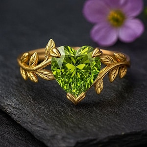 May include: A gold ring featuring a heart-shaped, faceted, light green gemstone. The ring's band is adorned with gold leaf detailing. The ring is set against a dark background, with a blurred purple flower in the background.