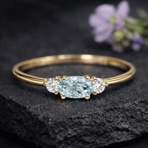 Könnte beinhalten: Ein goldener Ring mit einem ovalen, hellblauen Edelstein, flankiert von zwei kleineren, runden, weißen Edelsteinen. Der Ring liegt auf einer dunklen, strukturierten Oberfläche, mit einer verschwommenen lila Blume im Hintergrund.