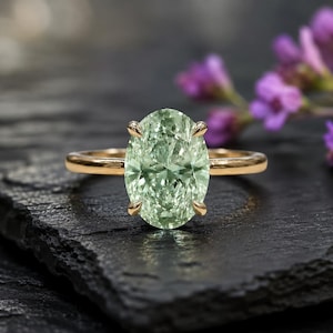 May include: A gold ring featuring a large, oval-cut, light green gemstone held in place by four prongs. The band is smooth and polished, showcasing a simple design that highlights the faceted, pale green stone as the central focal point.