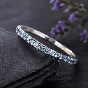 May include: A silver ring featuring a continuous row of small, pale blue gemstones. The ring is displayed on a dark, textured surface, with soft purple flowers in the background. The gemstones are evenly set around the ring's band.
