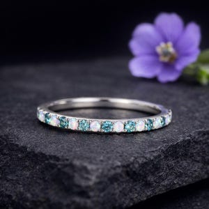 May include: A silver band ring featuring alternating white and teal-colored gemstones. The ring is set against a dark background, with a blurred purple flower in the background. The gemstones are round and evenly spaced around the band.