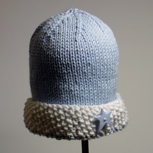 Pode incluir: Um chapéu de malha azul claro com uma borda branca e um botão de estrela azul.