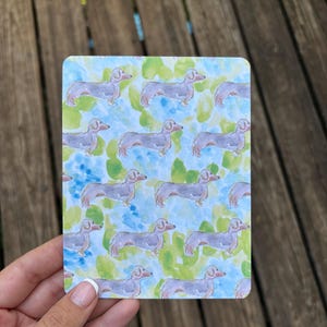 Puede incluir: Una tarjeta rectangular con un patrón repetido de perros salchicha. Los perros están representados en un estilo de acuarela con cuerpos grises y orejas rosas. El fondo tiene manchas de acuarela azules y verdes.