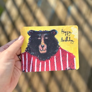 May include: A birthday card featuring a bear illustration. The bear has black fur, a red and white striped shirt, and a light brown snout. The card has a yellow background with the words "Happy birthday" written in black script.
