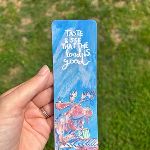 May include: A blue bookmark with the text "TASTE & SEE THAT THE lord IS good" in white script. The bookmark features a colorful illustration of a moose holding a mug. The background is a watercolor-style sky.