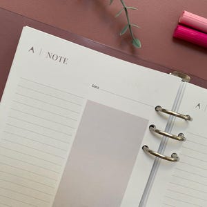 Può includere: Agenda aperta con pagine a righe e la parola "NOTE" stampata in alto. L'agenda è aperta su una pagina vuota con un rettangolo grigio chiaro. Sono visibili due pennarelli rosa e un rametto di verde.
