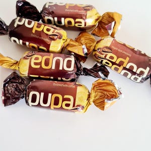 May include: Close-up of several individually wrapped candies. The wrappers are brown and gold, with the word "pupa" and "fednd" printed in white. The text "Enriched with coffee" is also visible.