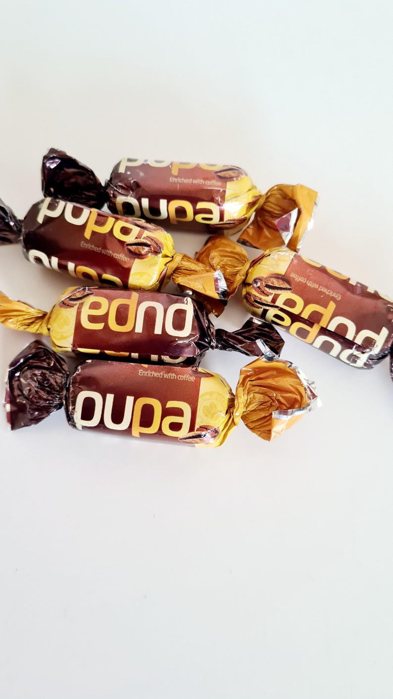 May include: Close-up of several individually wrapped candies. The wrappers are brown and gold, with the word "pupa" and "fednd" printed in white. The text "Enriched with coffee" is also visible.