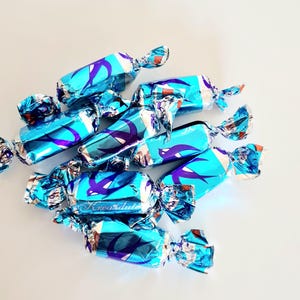 May include: A cluster of wrapped candies in shades of blue and silver, arranged on a white surface. The wrappers feature a repeating design in purple and the text "Krea dulte". The candies are individually wrapped.