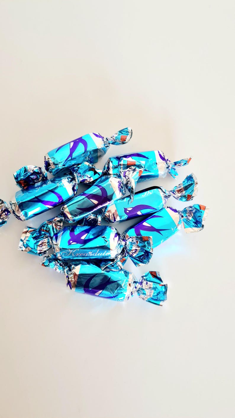 May include: A cluster of wrapped candies in shades of blue and silver, arranged on a white surface. The wrappers feature a repeating design in purple and the text "Krea dulte". The candies are individually wrapped.