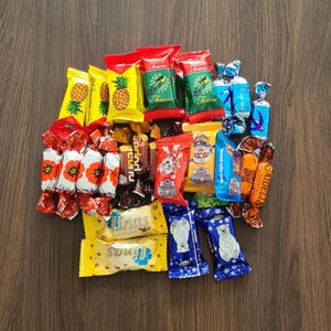May include: A collection of individually wrapped candies in various colors and designs. The assortment includes yellow pineapple-themed wrappers, red and green wrappers, and blue and white wrappers with polar bear illustrations. The candies are arranged on a wooden surface.