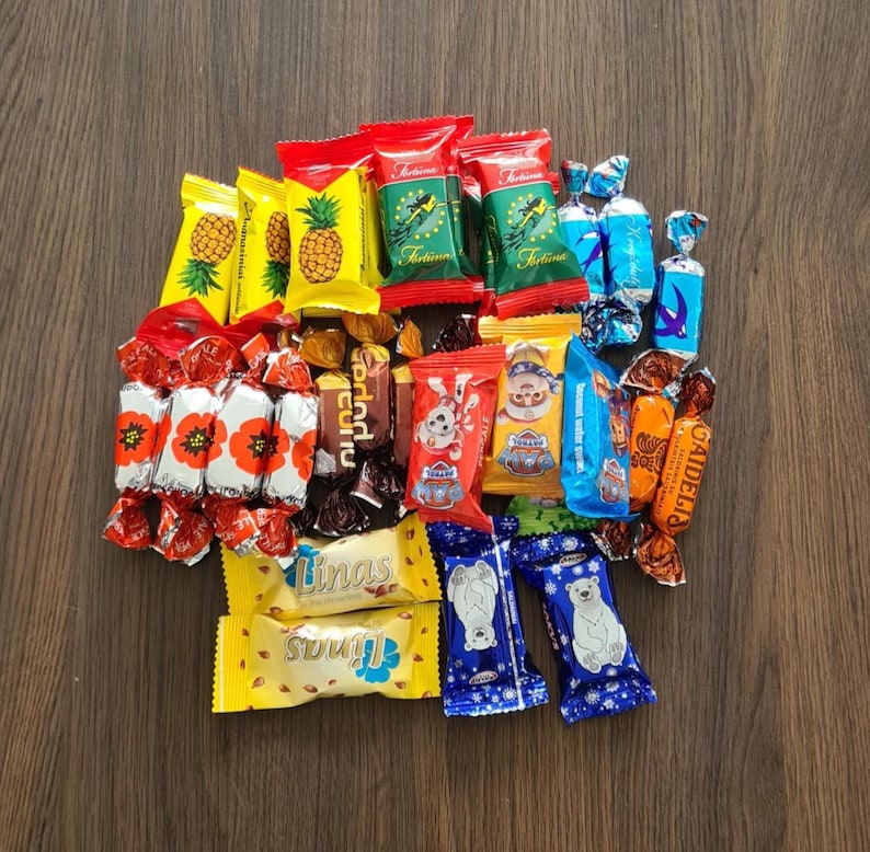May include: A collection of individually wrapped candies in various colors and designs. The assortment includes yellow pineapple-themed wrappers, red and green wrappers, and blue and white wrappers with polar bear illustrations. The candies are arranged on a wooden surface.