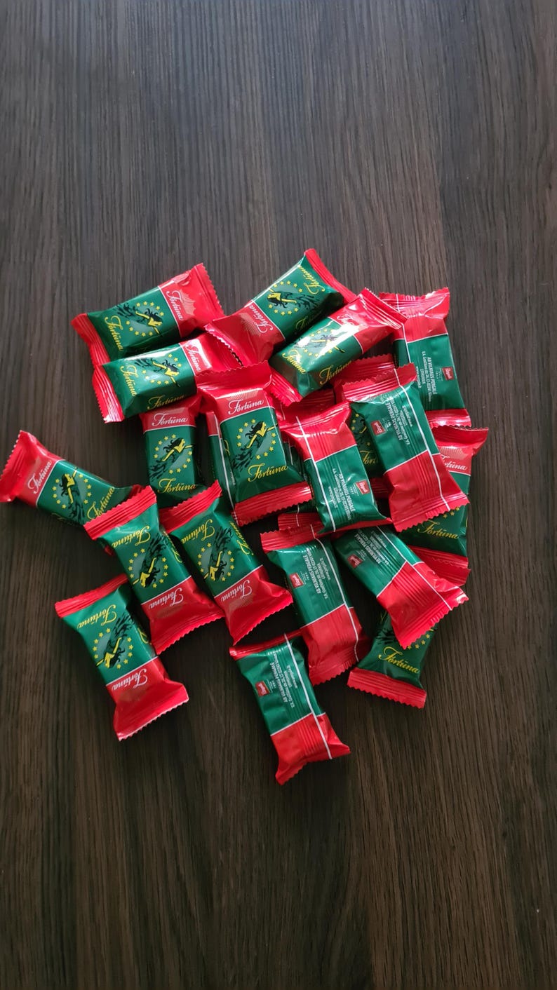 May include: A collection of individually wrapped candies with red and green packaging. The wrappers feature the word "Fortuna" in a script font, along with a graphic of a woman. The candies are scattered on a dark wooden surface.