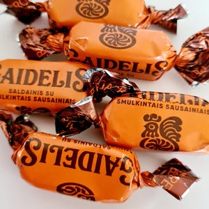 May include: Orange candy wrappers with the word "AIDELIS" printed in brown. The wrappers are twisted at the ends and feature a rooster graphic and text in a foreign language. The candies are arranged on a white surface.