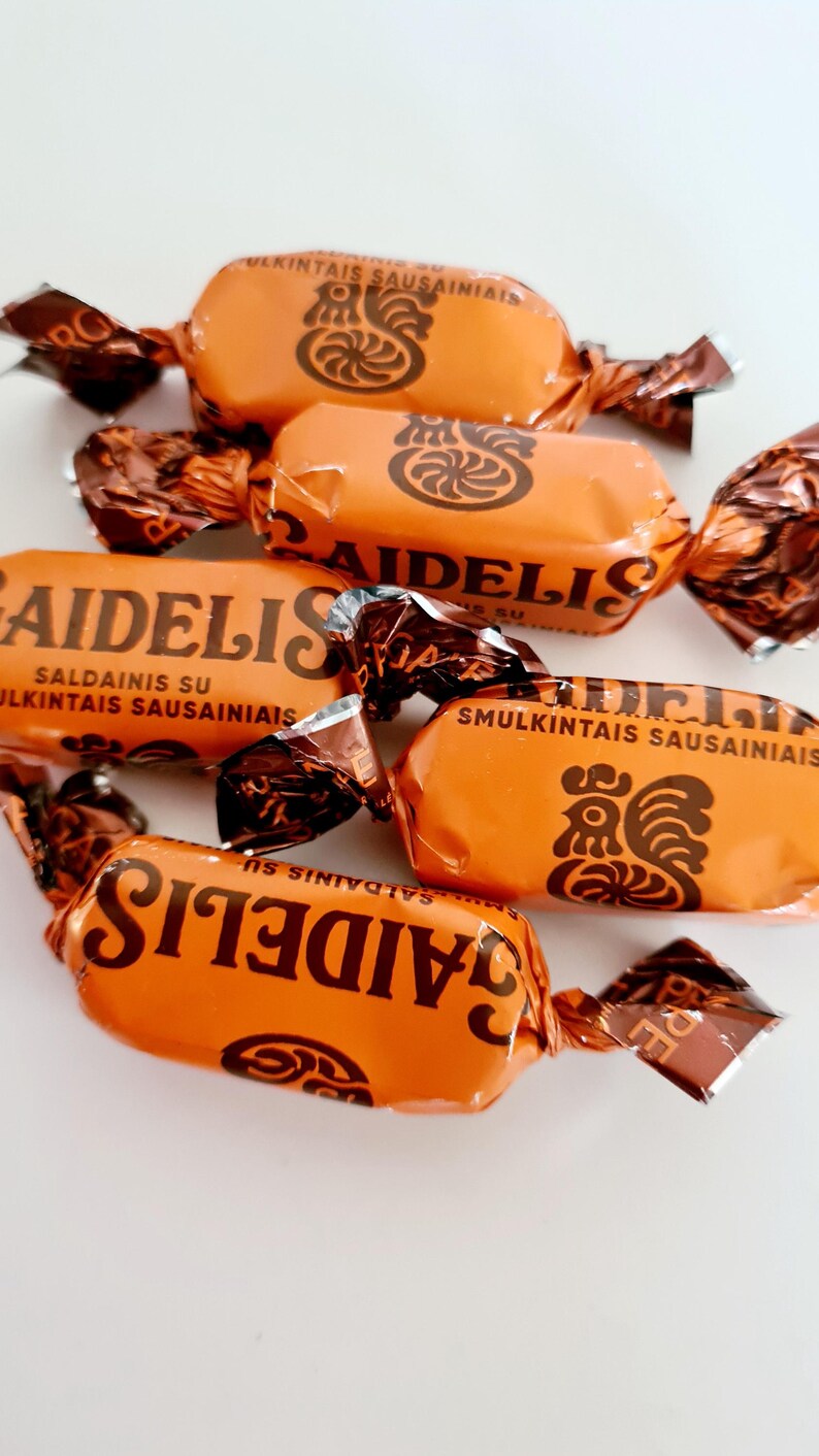 May include: Orange candy wrappers with the word "AIDELIS" printed in brown. The wrappers are twisted at the ends and feature a rooster graphic and text in a foreign language. The candies are arranged on a white surface.