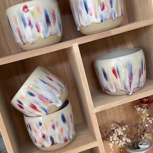 Könnte beinhalten: Eine Sammlung von Keramikbechern mit cremefarbener Basis und vertikalen, mehrfarbigen Tropfglasurmustern in Blau, Rot, Gelb und Pink. Die Becher sind in Holzregalen neben einer rosafarbenen Vase mit einer Zitronengrafik angeordnet.