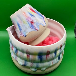 Könnte beinhalten: Ein Keramikbecher und eine passende Schale mit Ausguss, beide mit beigefarbenem, unglasiertem Boden und einem oberen Bereich mit weißer Glasur, der vertikale Streifen in Blau, Rot und Gelb aufweist. Das Innere der Schale ist hellrosa und enthält Füllmaterial.