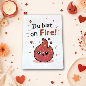 Puede incluir: Una tarjeta rectangular con acabado holográfico que presenta el texto "Du bist on Fire!" en letras negras y rojas. Debajo del texto hay una ilustración de dibujos animados de una llama roja sonriente con ojos grandes y estrellas.