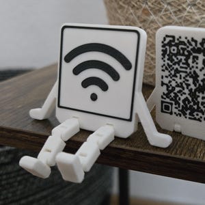 Könnte beinhalten: Zwei weiße, quadratische Figuren mit Armen und Beinen, die auf einer dunkelbraunen Oberfläche sitzen. Eine zeigt ein Wi-Fi-Symbol, die andere einen QR-Code. Die Figuren befinden sich neben einem geflochtenen Korb.
