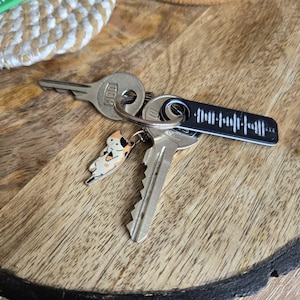 Könnte beinhalten: Ein Schlüsselbund auf einer Holzoberfläche. Die Schlüssel umfassen einen Schlüssel mit einem schwarzen Anhänger mit einem Schallwellenmuster, einen Schlüssel mit einem Katzenanhänger und zwei weitere Schlüssel.