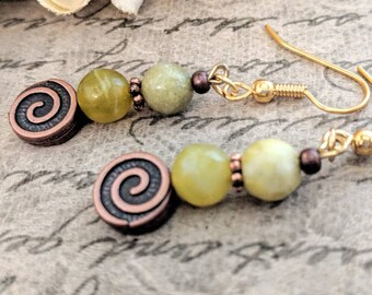 May include: A pair of dangle earrings featuring gold-tone ear wires, each with two round light green beads, a small dark metal spacer bead, and a circular dark copper-tone charm with a raised spiral design on the front surface.