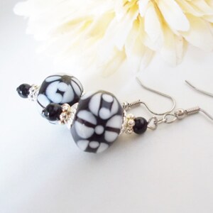 May include: Dangle earrings with black and white patterned beads and silver-tone accents. The earrings have a floral design and are suspended from silver-coloured ear wires. The background includes a soft, white flower.
