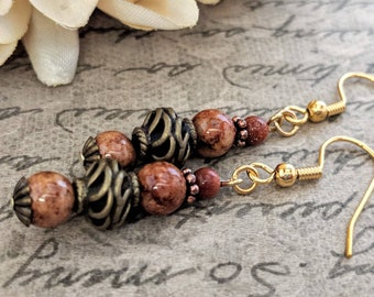 May include: Gold-tone dangle earrings featuring a combination of brown, bronze, and reddish-brown beads. The earrings have a unique design with filigree and textured beads, and gold-colored ear wires.