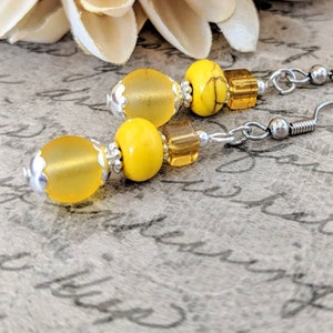 May include: Dangle earrings with yellow and silver beads. The earrings feature a combination of matte and glossy yellow beads, silver accents, and ear wires. The design incorporates round and square beads.