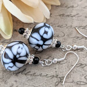 May include: Dangle earrings with large, round beads patterned in white and black. The earrings have silver-coloured metal accents and ear wires, with small black beads. The design is elegant and suitable for various occasions. The earrings are approximately 3 cm in length.