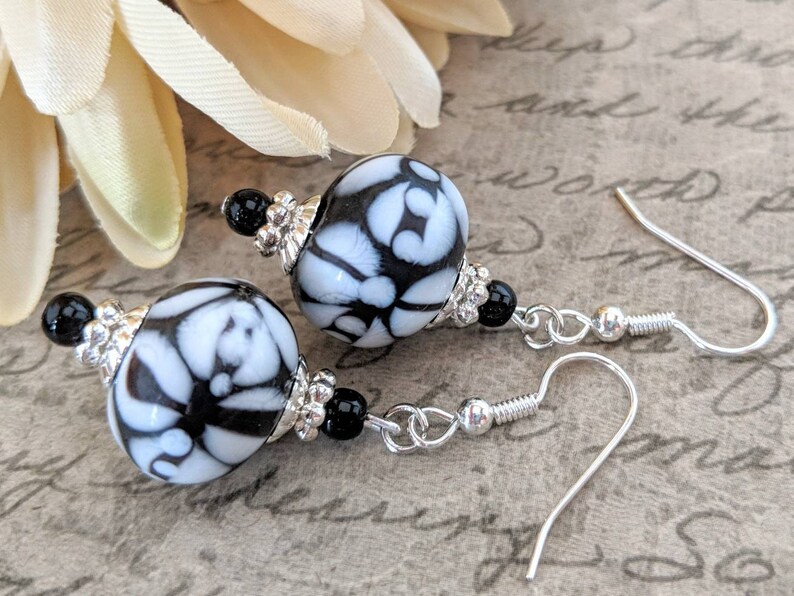 May include: Dangle earrings with large, round beads patterned in white and black. The earrings have silver-coloured metal accents and ear wires, with small black beads. The design is elegant and suitable for various occasions. The earrings are approximately 3 cm in length.