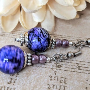 May include: A pair of drop earrings with large, round, marbled purple beads. The beads are accented with silver-toned metal and small, mauve-coloured beads. The earrings have silver-coloured ear wires, ideal for adding a touch of colour to any outfit.