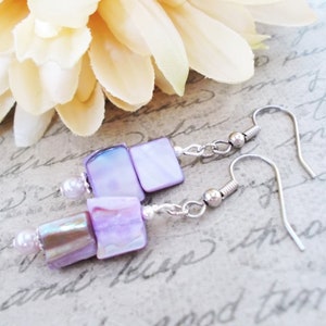 May include: Dangle earrings with iridescent lavender and purple square beads, linked by silver chains and small beads. The earrings have silver fish hook closures. The image shows the earrings near a cream-coloured flower.