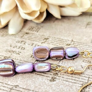 May include: Two pairs of earrings with iridescent, lavender-coloured, square beads and gold-coloured hooks. The beads have a pearlescent sheen. The background includes sheet music and a soft, cream-coloured flower. The earrings are a delicate piece of jewellery.