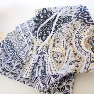 May include: A pile of folded fabric napkins featuring diverse patterns. The designs include geometric shapes, paisley, and chevron patterns in black, grey, white, and gold. A white ribbon is visible.