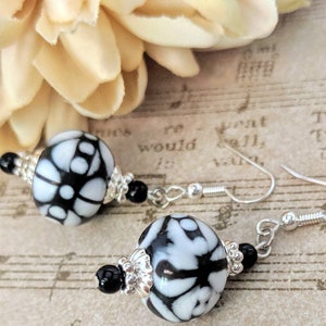 May include: Dangle earrings with large, round beads in a black and white floral design. The earrings have silver-coloured hooks and are accented with small black beads and silver-toned decorative elements. A cream-coloured flower is in the background.