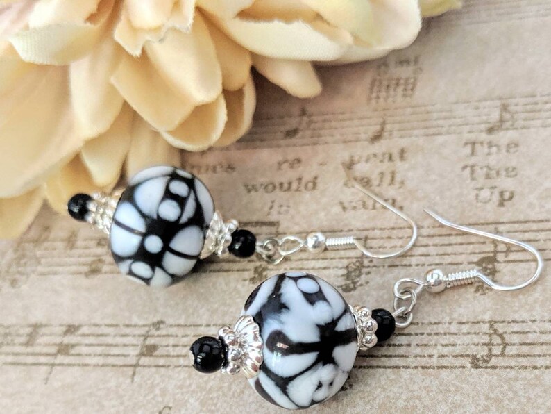 May include: Dangle earrings with large, round beads in a black and white floral design. The earrings have silver-coloured hooks and are accented with small black beads and silver-toned decorative elements. A cream-coloured flower is in the background.
