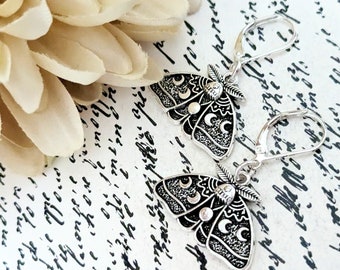 May include: A pair of silver leverback earrings, each with a detailed moth design. The moths have crescent moon and dot accents, with textured wings. The earrings have a vintage style. The background includes a cream flower and script.