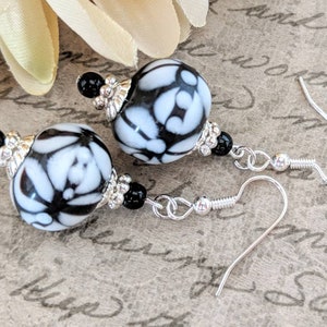 May include: Dangle earrings with large, round beads patterned in black and white floral designs. The earrings have silver-coloured hooks and accents, with small black beads and silver-coloured bead caps.