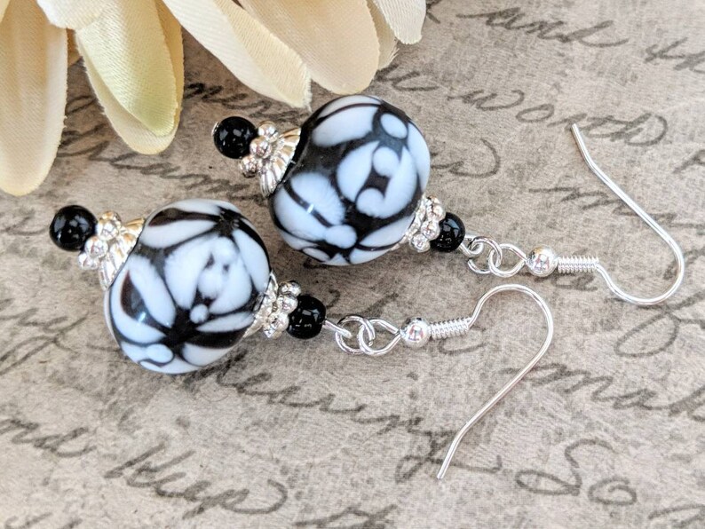 May include: Dangle earrings with large, round beads patterned in black and white floral designs. The earrings have silver-coloured hooks and accents, with small black beads and silver-coloured bead caps.