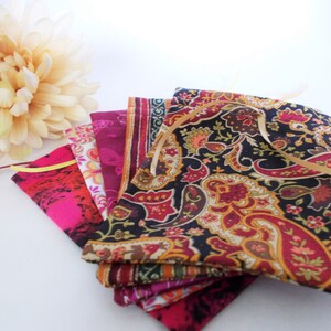 Op de afbeelding: Een verzameling kleurrijke stoffen zakjes met gouden linten, met verschillende patronen, waaronder paisley en bloemmotieven. De zakjes zijn in tinten roze, rood en bordeaux, met een cr&egrave;me-kleurige bloem op de achtergrond.
