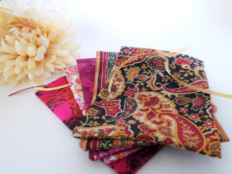 Op de afbeelding: Een verzameling kleurrijke stoffen zakjes met gouden linten, met verschillende patronen, waaronder paisley en bloemmotieven. De zakjes zijn in tinten roze, rood en bordeaux, met een cr&egrave;me-kleurige bloem op de achtergrond.