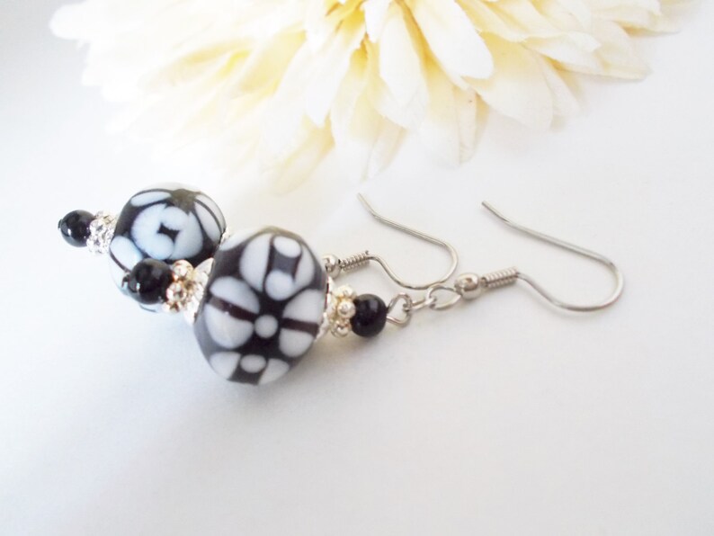 May include: Dangle earrings with black and white patterned beads and silver-tone accents. The earrings have a floral design and are suspended from silver-coloured ear wires. The background includes a soft, white flower.