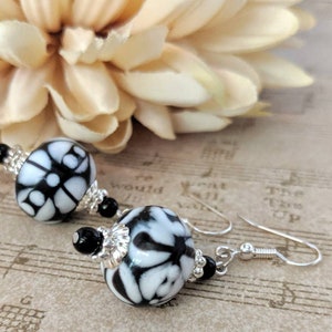 May include: A pair of drop earrings with round beads patterned in black and white. The beads have a floral design and are accented with silver-coloured metal and black beads. The earrings hang from silver-coloured ear wires.