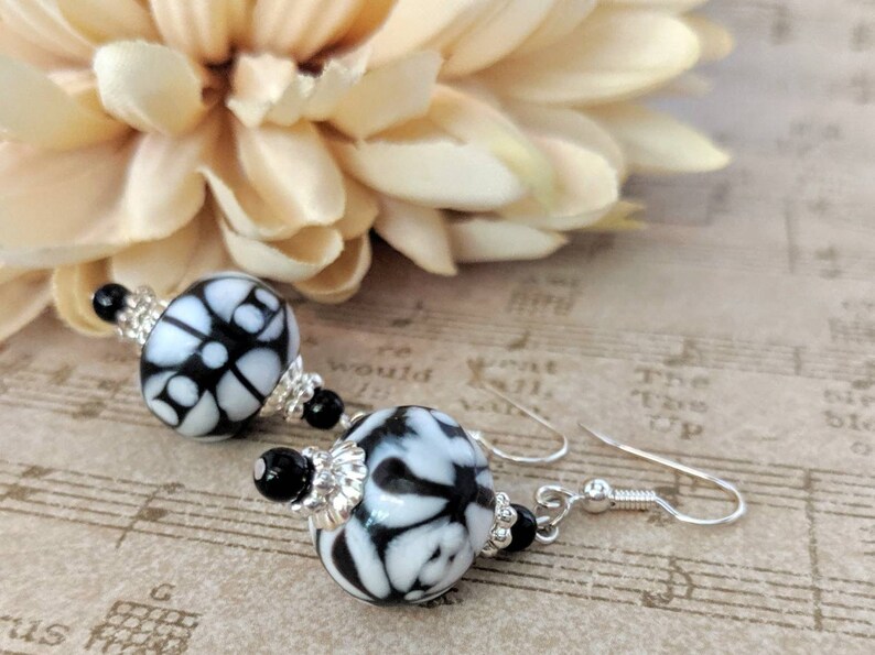 May include: A pair of drop earrings with round beads patterned in black and white. The beads have a floral design and are accented with silver-coloured metal and black beads. The earrings hang from silver-coloured ear wires.