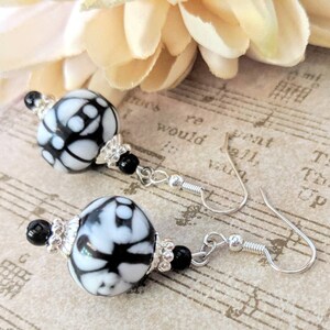May include: Dangle earrings with large, round beads in a black and white pattern. The earrings have silver-coloured hooks and are accented with small black beads and silver-coloured decorative elements. The background includes a flower and sheet music.