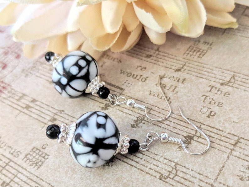 May include: Dangle earrings with large, round beads in a black and white pattern. The earrings have silver-coloured hooks and are accented with small black beads and silver-coloured decorative elements. The background includes a flower and sheet music.