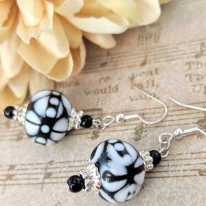 May include: Dangle earrings with large, round beads patterned with white and black floral designs. The earrings have silver-coloured hooks, small black beads, and silver-coloured bead caps. A beige flower is in the background.