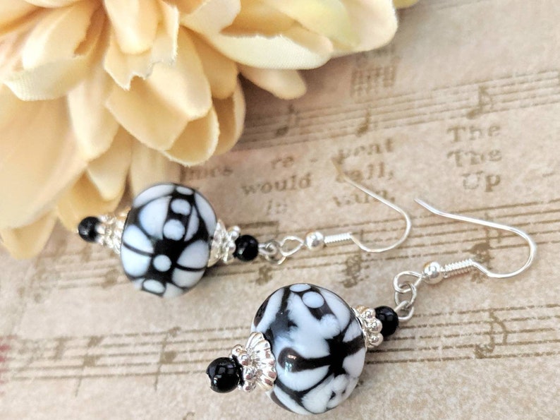 May include: Dangle earrings with large, round beads patterned with white and black floral designs. The earrings have silver-coloured hooks, small black beads, and silver-coloured bead caps. A beige flower is in the background.