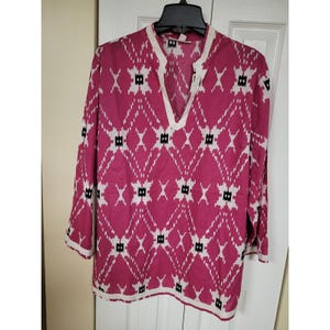 May include: A long-sleeved tunic in a vibrant fuchsia hue, adorned with a geometric pattern in white and black. The tunic features a V-neckline and white trim along the collar and cuffs. The garment is hanging on a black hanger.