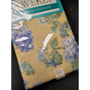 May include: A rectangular tablecloth with a floral pattern of blue and green hydrangeas on a tan background. The tablecloth is 60" x 84" and is packaged in clear plastic. The package reads "Easy Care Tablecloth".